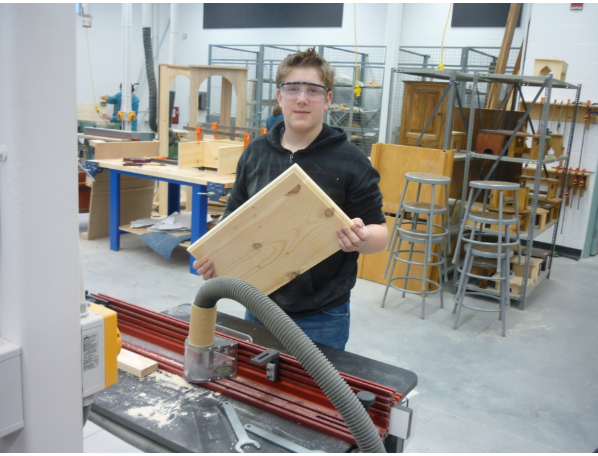 Student stands in woodshop by a machine holding a rectangular piece of wood